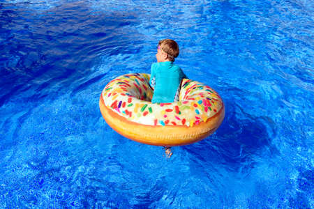 A boy on his back cools off on an inflatable float in a pool, wearing a bathing suit to protect...