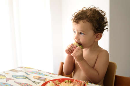 A young girl sitting in her high chair grabs a piece of vegetables and eats it by herself.