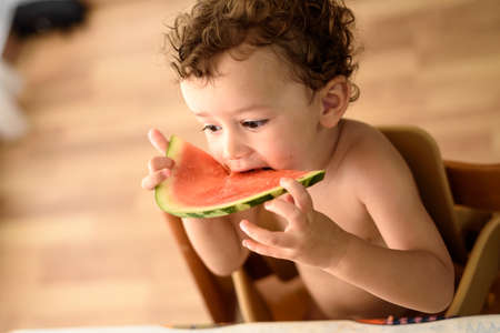 Curly-haired baby girl eats fruit sitting indoors at home in summer.の写真素材