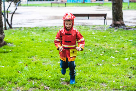 Boy dressed as a fireman with red raincoat splashes on the grass of a park a rainy day.の写真素材