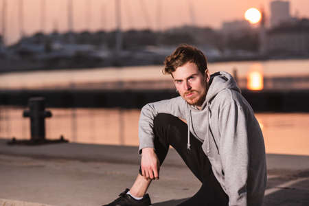 Portrait of young red-haired man with gray sweatshirt sitting on the jetty of a harbor at sunset.の写真素材