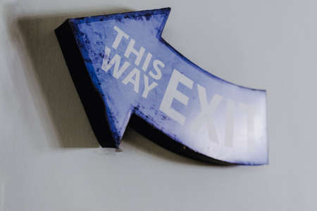 This Way Exit written on a decorative blue wooden arrow hanging on a wall.