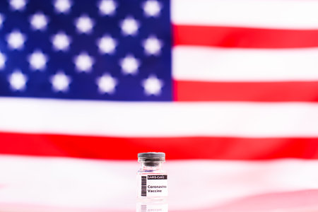 Airtight transparent vial with the new vaccine against the covid disease caused by coronavirus, with the background of the American flag out of focus.の写真素材