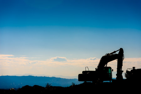 Backlit silhouette of an excavator on top of a hill with unfocused sky background.の写真素材