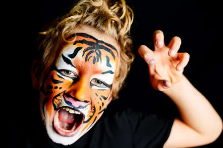 A growling boy with his face painted in the striking colors of a tiger, isolated on a black studio...