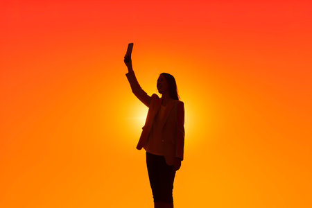 A young woman takes a selfie, raising her cell phone, against the sun in silhouette.の素材