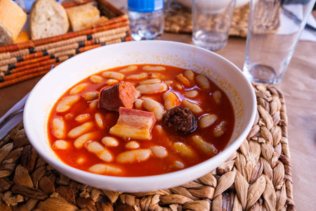 A bowl filled with Fabada Asturiana, a traditional Spanish bean and meat soup, sits on a wooden table. The soup is hearty and flavorful, containing tender pieces of meat and creamy beans.の写真素材