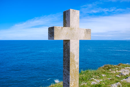A Christian cross stands prominently on a hill, overlooking the vast expanse of the ocean. The cross is silhouetted against the horizon, with waves crashing in the distance.の写真素材