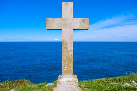 A cross stands tall on a hill, casting a shadow against the backdrop of the vast ocean with waves below. The scene exudes a sense of solemnity and reverence.の写真素材