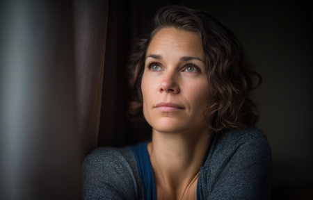A woman with curly hair is lost in thought while gazing out of a window. The soft light filters through the curtains, creating a peaceful atmosphere that evokes emotions of calmness and reflection.の素材