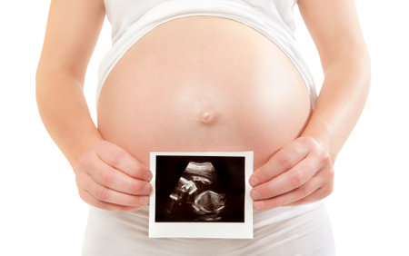 Pregnant woman holding ultrasound examination picture in front of her round belly, isolated on white...