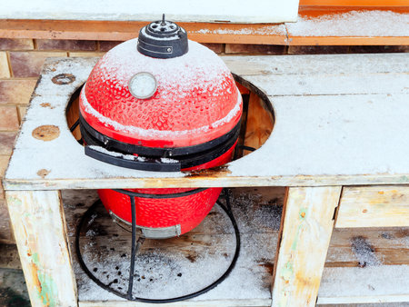 Red ceramic BBQ grill lightly dusted with snow in the hole of the wood tableの写真素材