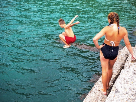 A girl stands on a concrete pier and watches as a boy jumps into the muddy sea with bent legsの写真素材