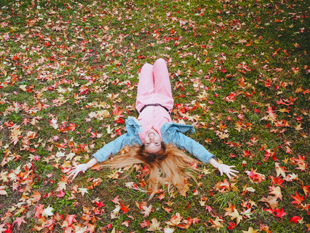 Red-haired smiling girl lies with her arms spread on a meadow strewn with autumn maple leaves