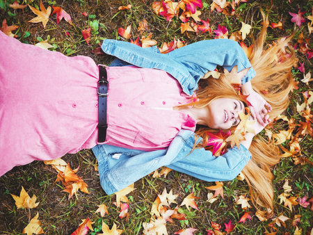Red-haired cheerful girl lies in a meadow covered with maple leaves with her palms folded above her headの写真素材