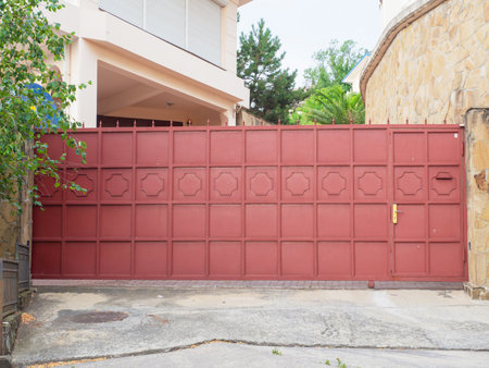 A red iron fence with a wicket gate, geometric pattern and sharp spiers encloses a modern residential building and a courtyard with various green treesの写真素材