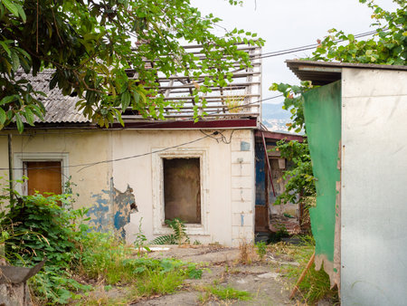 An old abandoned one-story house with a destroyed roof and broken windows and grass growing through the concrete flooring in the yardの写真素材