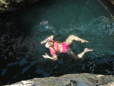 A girl swims in a frog pose in a turquoise river lit by the sun