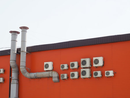 A fragment of an orange building with many air conditioners hanging on the wall and two ventilation...