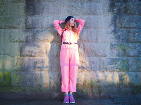 A girl in a pink jumpsuit stands holding her hands behind her head near a stone wall in the sunlight