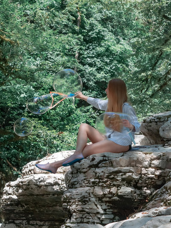 The girl blows big soap bubbles while sitting on the canyons on the background of the forest