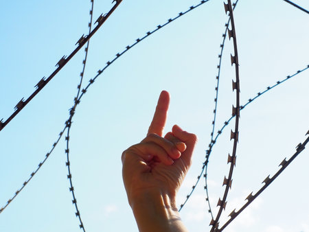 Hand shows middle finger against the background of barbed wire and blue skyの写真素材