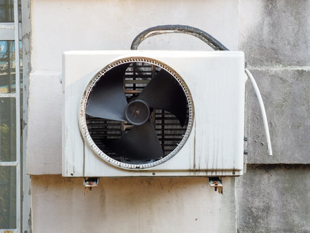 An air conditioner unit with a fan and a drain pipe hangs on the wall outside.の写真素材