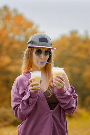 A girl in a purple shirt with purple lips drinks two milkshakes from tubules at the same time on an autumn dayの写真素材