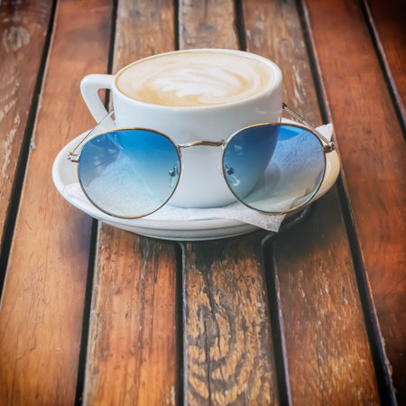 White cup of cappuccino with sunglasses on a saucer in front of it stands on a wooden surface
