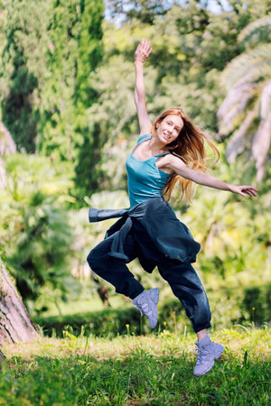 Red-haired hip-hop girl jumping with her arms spread on the lawn in a green park on a sunny summer dayの写真素材