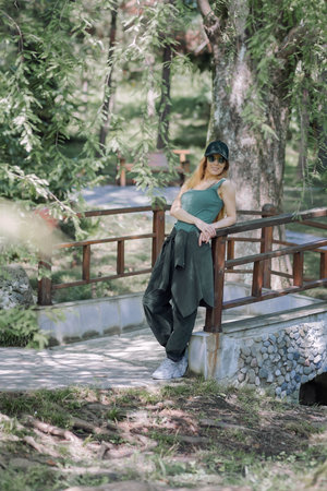 Smiling sporty girl in green outfit stands near the bridge leaning her elbow on the railing in a summer parkの写真素材
