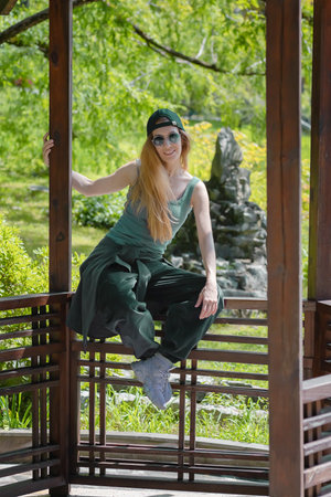 Smiling woman in emerald hip-hop outfit sits on the side of a wooden gazebo in a summer green parkの写真素材
