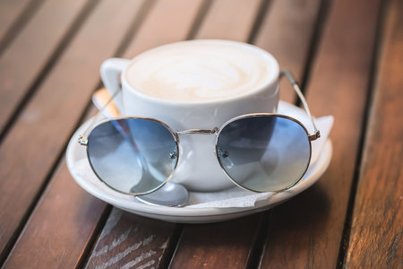 White cup of cappuccino with sunglasses stands on a wooden surfaceの写真素材