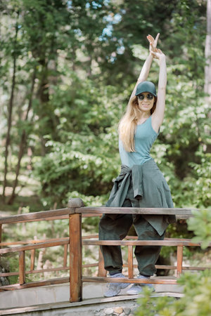 Modern blonde woman in hip-hop outfit stands with her arms outstretched above her head on a bridge with wooden railing in a green parkの写真素材