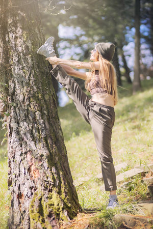 A woman in a sports outfit does a stretch by throwing her foot on the trunk of a tree standing on a hill in a sunny park.の写真素材