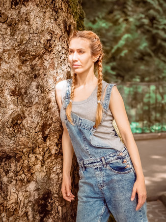 Lady with pigtails in denim overalls is leaning on the trunk of a massive tree in the parkの写真素材