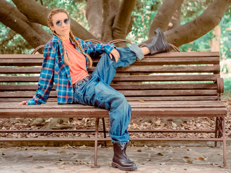 A girl with pigtails in a plaid shirt with oversize jeans and rough shoes sits on a bench under a sprawling tree.の写真素材