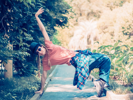A lady with pigtails in a hat and streetwear stands on her toes, leaning on her hand on a path in a green park.の写真素材