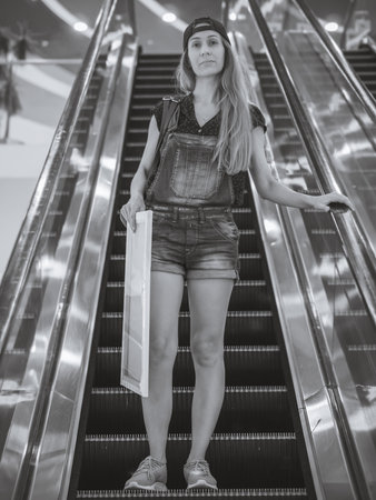 A girl in a short denim overalls rides down the escalator holding a canvas on a stretcher in her hand. Black and white photoの写真素材