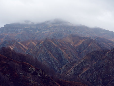 Rocky mountain ranges with dry yellow grass, bare autumn trees and overhanging clouds. Mountains of...