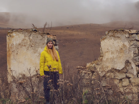 A woman stands near the wall of a ruined house in tall dry grass against the backdrop of autumn hills with grazing cows and foggy haze in the distanceの写真素材