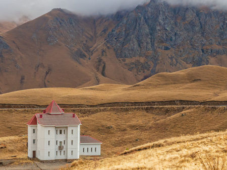 High in the autumn mountains of the Caucasus, where the clouds lie on the peaks, there is a white house with narrow windows and a red roof.の写真素材