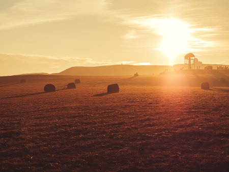 A golden field with haystacks on a mountain peak with a gazebo and tourists in the distance in a twilight haze and in the rays of the setting sun. Soft focus.の写真素材