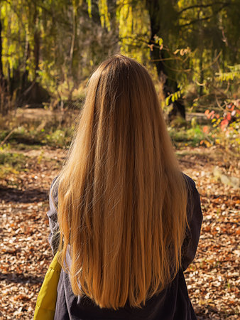A fair-haired woman with long flowing hair stands on a sunny day in an autumn park. Back view. The...