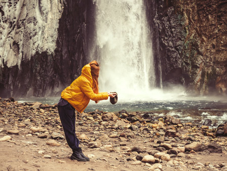 A woman stands on the bank of a river against the backdrop of a stormy waterfall, filling her hat with water from it. Optical illusionの写真素材