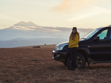 A woman stands near an SUV in a high-mountain field against the backdrop of Elbrusの写真素材