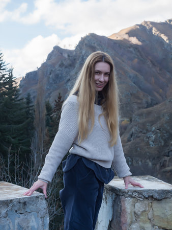 Smiling woman stands between stone old structures against the background of autumn trees and steep...