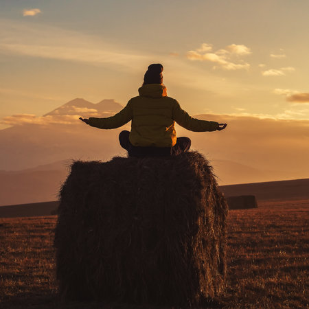 A woman sits on a haystack spreading her palms to the sides against the backdrop of Elbrus in the clouds at sunset. View from the backの写真素材