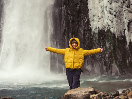 Smiling woman showing thumbs up against the background of an icy waterfall in a rocky areaの写真素材