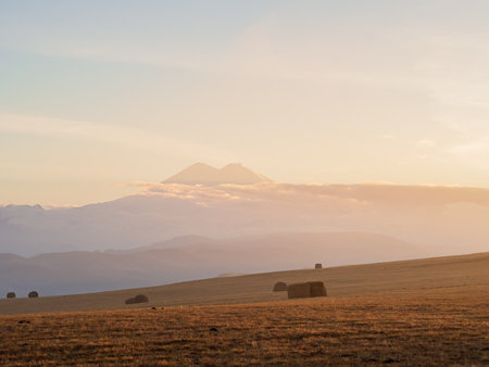 Elbrus in the distance against the twilight sky among clouds, mountain ranges and golden fieldsの写真素材